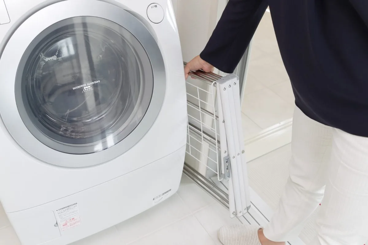 洗濯機の横にコンパクトに折り畳んだWY物干しスタンドを収納する女性
