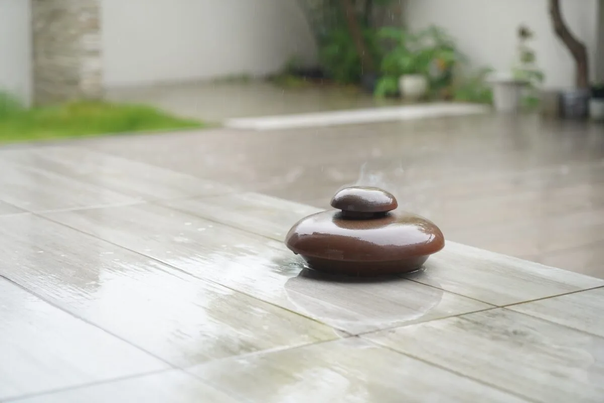雨の日の屋外で煙を上げる備前焼蚊遣り、濡れたタイルの上に置かれた耐雨仕様の蓋付き設計