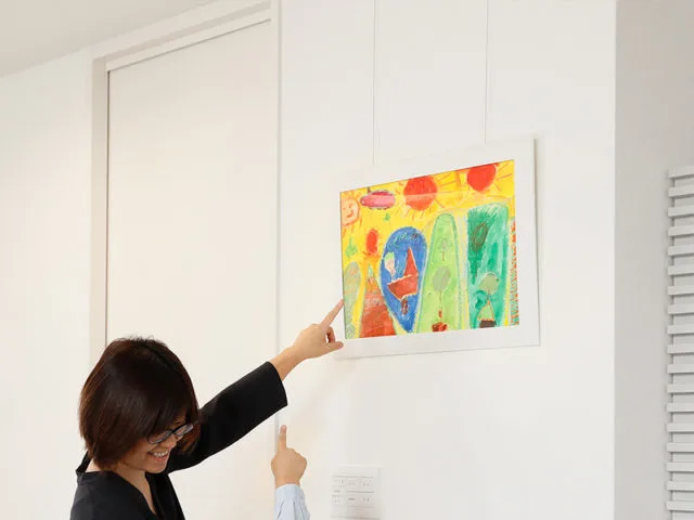 女性が壁にワイヤーで吊るした額縁入りの子供の絵を指差して紹介している