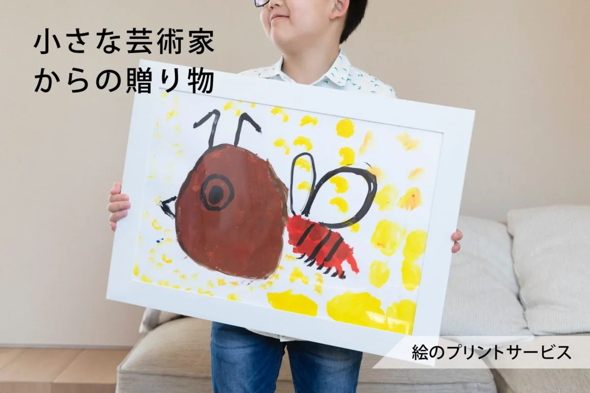 子どもがミツバチの絵を印刷したホワイトフレームを抱えて微笑む様子。「絵のプリントサービス」の文字入り