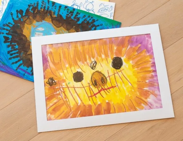 白いペーパーフレームに子どもの水彩画を飾った様子、木目の床に複数の絵が並ぶ