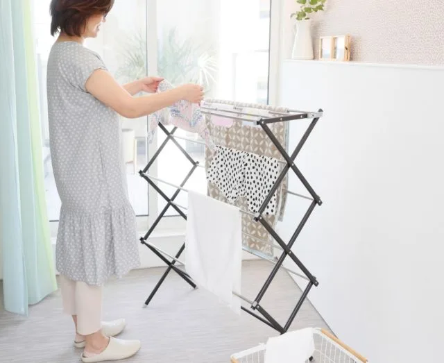 女性がX型折りたたみ物干しスタンドに洗濯物を干している室内シーン
